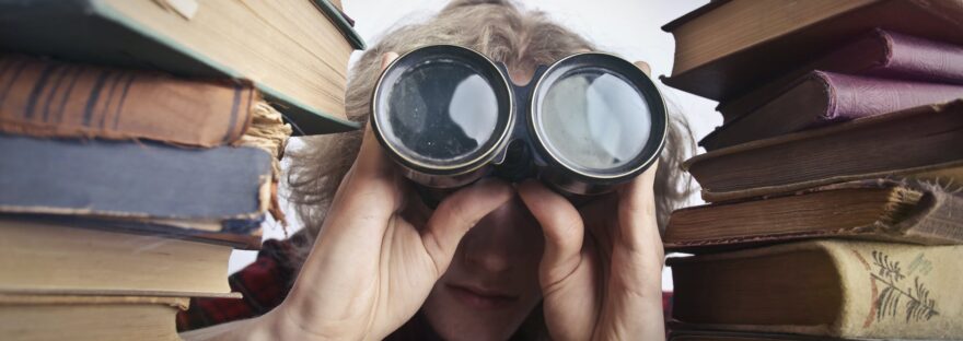 anonymous person with binoculars looking through stacked books