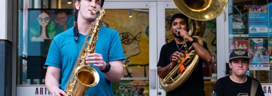 three man playing drums and wind instruments in front of store